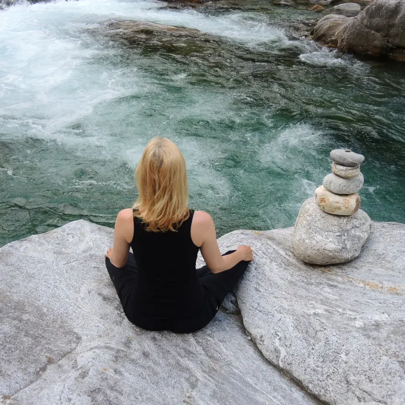 Frau sitzt still auf einem Felsen am Gebirgsfluss, daneben ein kleines Steinmännchen